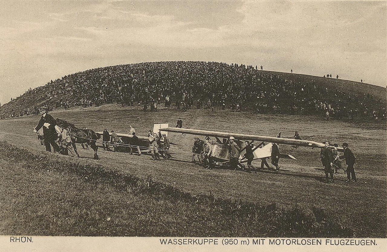 Schwaz-weiß-Foto: Viele Menschen stehen auf einer Wiese, in der Mitte ein Segelflugzeug, das von einem Pferd gezogen wird.